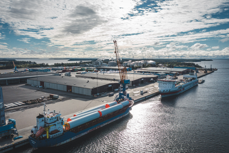 Fotografisk drönarbild över en hamn med klart, blått vatten. Två fartyg ligger intill kajen, stilla och tydliga i bilden. Himlen är klar med några lätta, vita moln som breder ut sig mjukt ovanför hamnområdet. Bilden har en lugn och fridfull atmosfär.