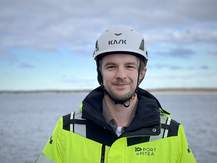 En man står framför kameran och ler. Han tittar in i kameran. På sig har han en hjälm och en varselväst. I bakgrunden syns havet.