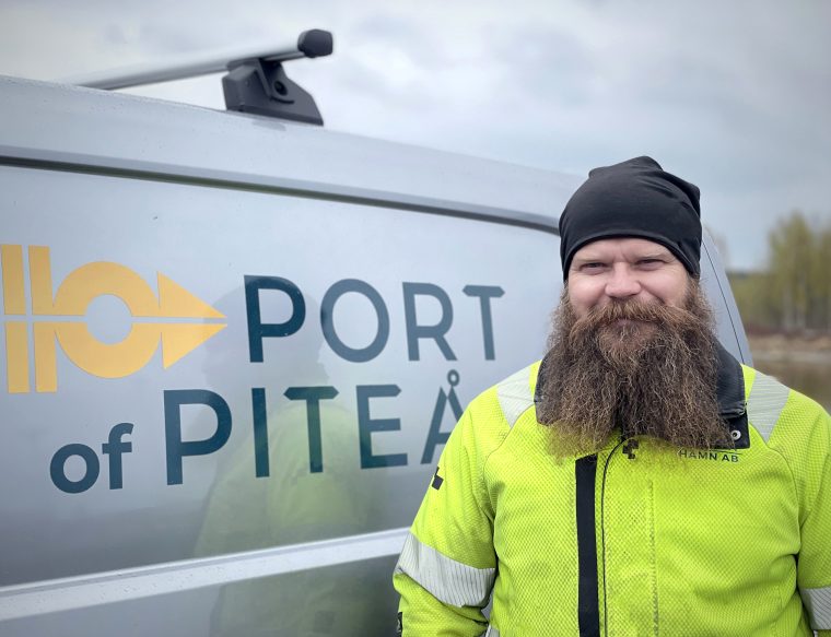 En man med skägg står framför en grå arbetsbil med logotypen 'Port of Piteå' på dörren. I bakgrunden syns en gråmulen himmel.