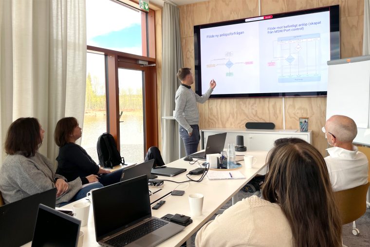 Bilden visar hur fyra personer sitter vid ett avlångt konferensbord. Längst fram står en man i jeans och grå tröja. Han håller i en utbildning och pekar med ena handen mot skärmen. På bordet står uppfällda bärbara datorer. Bilden ger ett lugnt intryck.