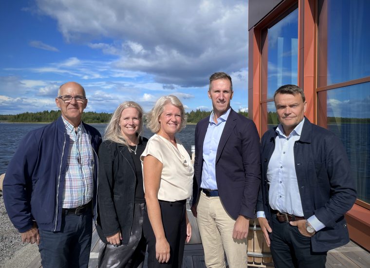 Fem personer står utomhus framför Piteå Hamns kontor, som är rostrött. I bakgrunden syns havet, en blå himmel med vackra moln och det ser soligt men lite blåsigt ut.