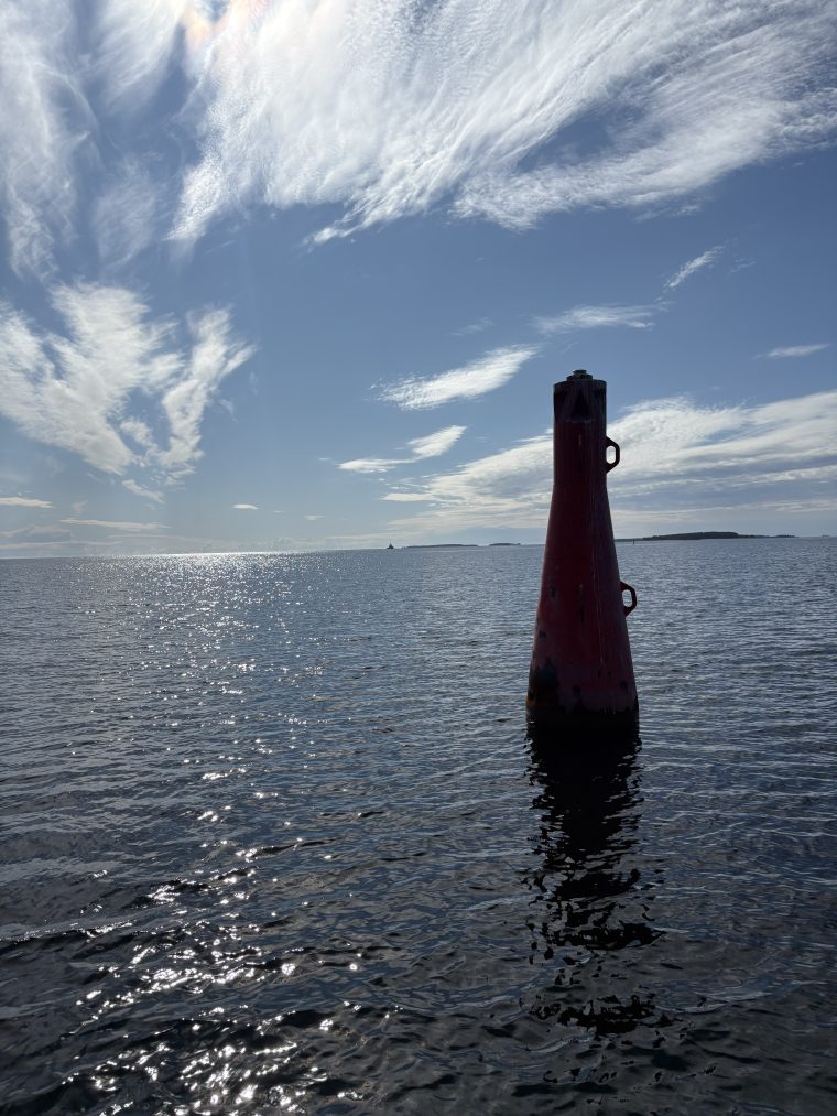 En röd lysprick reser sig ur vattnet, omgiven av öppet hav. Solen står lågt och lyser bakifrån, så bojens konturer framträder mörka mot det glittrande motljuset.