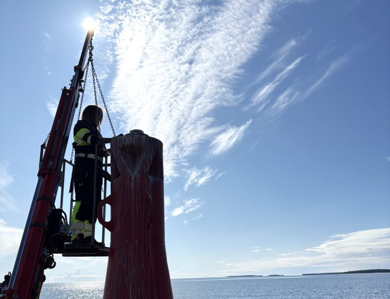 En drifttekniker står högt upp på en smal ställning intill en lysboj. Driftteknikern bär flytväst och arbetar koncentrerat med att justera utrustningen, medan havet breder ut sig i bakgrunden.