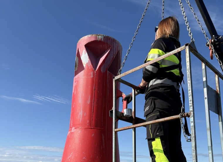 En drifttekniker står högt upp på en smal ställning intill en lysboj. Driftteknikern bär flytväst och arbetar koncentrerat med att justera utrustningen, medan havet breder ut sig i bakgrunden.