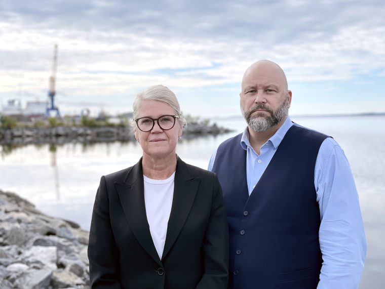 En man och en kvinna står bredvid varandra och ser rakt in i kameran. De är klädda propert men avslappnat, mannen i skjorta och kvinnan i en stilren klädsel. I bakgrunden syns havet och himlen, och hela bilden går i blå toner som ger ett lugnt, harmoniskt intryck.