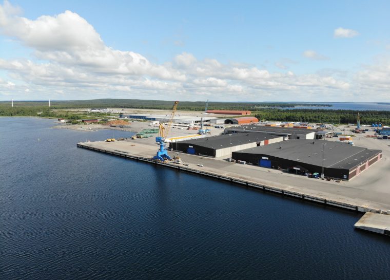 Drönarfoto över en lång kaj med två stora magasin och rymliga logistikytor. En blå kran syns vid kajen under klar blå himmel en stilla dag.