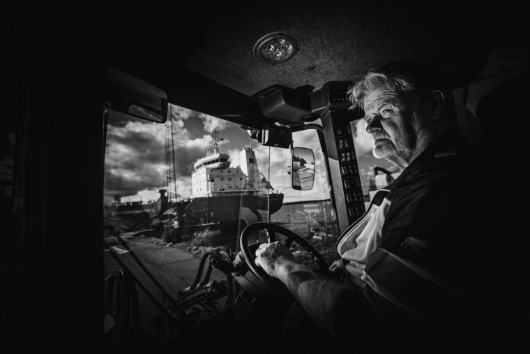 Svartvit bild med en man i förarhytten på en traktor. Hans ansikte är upplyst av en blixt, och i bakgrunden syns ett fartyg.