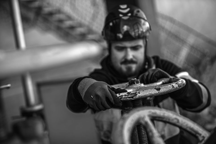 Svartvit närbild på en man i varselkläder och hjälm som manövrerar en kran. Hans blick är koncentrerad och visar fullt fokus på arbetet.