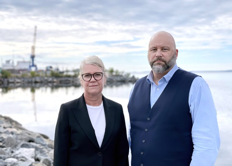 En man och en kvinna står bredvid varandra och ser rakt in i kameran. De är klädda propert men avslappnat, mannen i skjorta och kvinnan i en stilren klädsel. I bakgrunden syns havet och himlen, och hela bilden går i blå toner som ger ett lugnt, harmoniskt intryck.